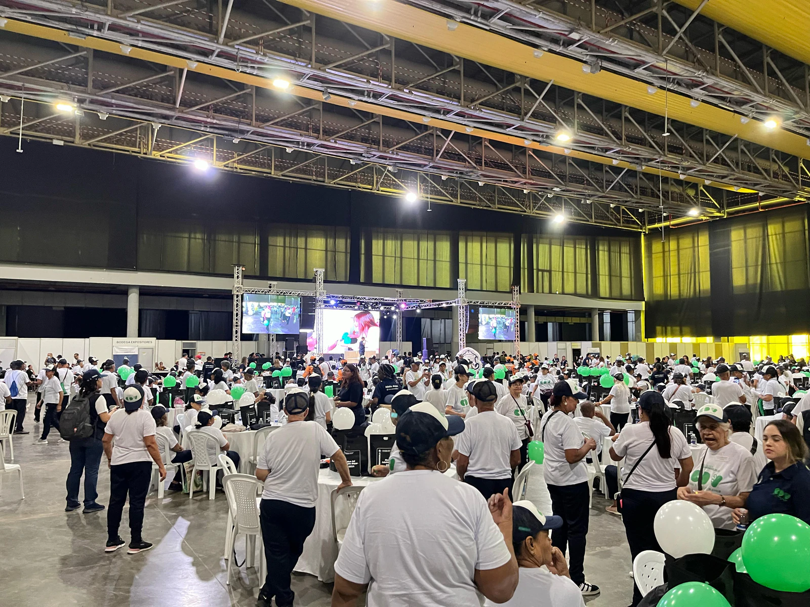 Encuentro en un salón con asistentes, pantallas y globos verdes y blancos